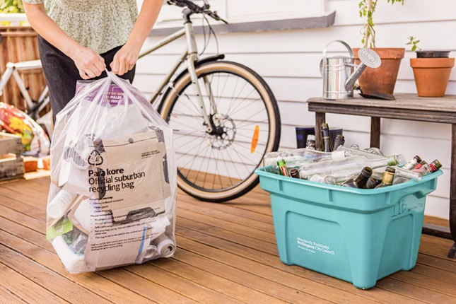 A large clear bag filled with recycling, being tied up by a person who is only visible waist down, standing on a deck with potted plants, a bicycle and a green recycling bin filled with glass.